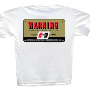 Op de afbeelding: Wit T-shirt met een gouden en rood waarschuwingsbord met de tekst "WARNING THIS CAR EQUIPPED WITH FLOOR HURST SHIFT SHIFT AS HARD AS YOU PLEASE BUT DON'T BREAK YOUR ARM"