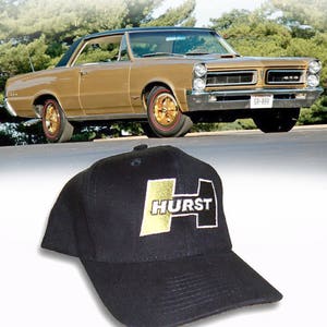 May include: A black baseball cap featuring a prominent embroidered logo on the front panel. The logo consists of a large stylised letter H in gold and black, with the word HURST printed in white capital letters across the centre of the design.