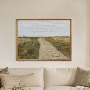May include: A rectangular landscape painting in a gold frame hanging above a cream-colored sofa. The artwork depicts a dirt path through a grassy field under a cloudy sky, featuring the text: You will show me the path of life; in your presence is fullness of joy; at your right hand are pleasures forevermore. Psalm 16:11.