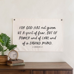 May include: A white canvas banner with black text that reads "FOR GOD HAS not given US A spirit of fear, BUT OF POWER and of LOVE and of a SOUND MIND. 2 TIMOTHY 1:7". The banner is hanging on a white wall above a wooden dresser.