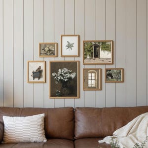 May include: A brown leather sofa with a white striped pillow and a cream-colored throw blanket. Above the sofa, a gallery wall of framed botanical and landscape prints hangs against a gray paneled wall. A gold side table holds a candle and a vase.