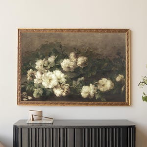 Framed vintage floral painting of white roses with green foliage, hanging above a modern black cabinet in a neutral-toned living room. The artwork features an ornate gold frame and a muted, antique-style background, complementing the minimal, elegant decor.