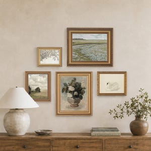 May include: A wooden console table with three drawers and a lower shelf holding a bowl, a lamp with a white shade, and a vase with greenery. Above the table, five framed art prints are arranged on the wall, featuring landscapes, a swan, and flowers.