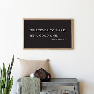 May include: A framed black print with the quote "WHATEVER YOU ARE. BE A GOOD ONE." by Abraham Lincoln. The print hangs above a metal bench with a pillow, baseball glove, baseball, and a green knitted throw. A black wire basket holds a football and a soccer ball.