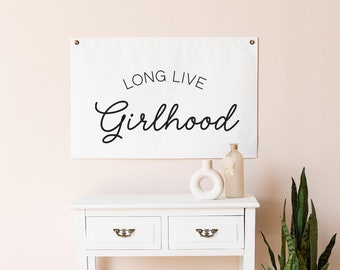 Op de afbeelding: Een wit rechthoekig wandbanner met de zwarte tekst "LONG LIVE Girlhood" in een combinatie van schreefloze en scriptlettertypes. Het banner is bevestigd met twee metalen ringen in de bovenhoeken tegen een lichtroze muur boven een witte tafel.