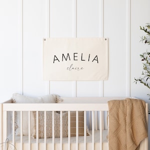 May include: A white canvas banner with black text that reads "AMELIA claire" hanging on a white wall above a white crib with a tan blanket and pillows.