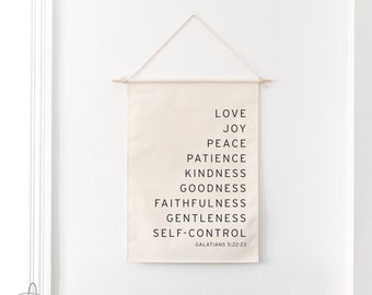 May include: A white canvas wall hanging with black text listing the fruits of the spirit: love, joy, peace, patience, kindness, goodness, faithfulness, gentleness, and self-control. The text also includes the scripture reference Galatians 5:22-23.