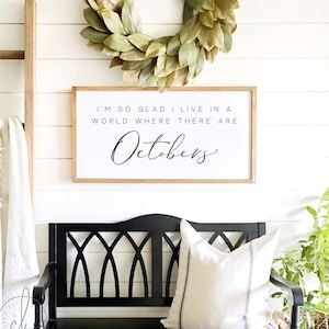 May include: A wooden sign with the text "I'm so glad I live in a world where there are Octobers." The sign is hanging on a white wall with a wreath above it. A black bench with a white pillow is in the foreground.
