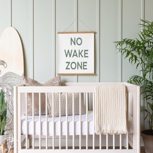 May include: A white crib with a white mattress, two pillows, and a cream-colored blanket. A wooden sign with the text "NO WAKE ZONE" hangs above the crib. A wooden surfboard and a potted plant are also in the room.