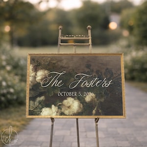 May include: A rectangular sign with a gold frame resting on a metal easel. The sign features a dark floral background with white flowers and the white text "The Fosters" in a script font, followed by the date "OCTOBER 5, 2026" in a sans-serif font.