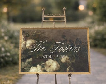 Könnte beinhalten: Ein rechteckiges Schild mit goldenem Rahmen auf einer Metallstaffelei. Das Schild zeigt einen dunklen floralen Hintergrund mit weißen Blumen und den weißen Schriftzug "The Fosters" in Schreibschrift, gefolgt vom Datum "OCTOBER 5, 2026" in serifenloser Schrift.