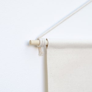 May include: A blank canvas wall hanging with a natural wooden dowel and a cream-colored rope for hanging. The canvas is a light beige color, and the rope is knotted around the dowel. The wall hanging is displayed against a white wall.