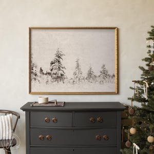 May include: A framed black and white print of a snowy forest scene. The artwork is in a gold frame and is displayed above a dark gray dresser. A Christmas tree with ornaments is to the right.
