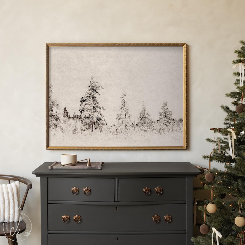 May include: A framed black and white print of a snowy forest scene. The artwork is in a gold frame and is displayed above a dark gray dresser. A Christmas tree with ornaments is to the right.