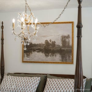 May include: A crystal chandelier with four lights hangs above a bed. A landscape painting with a gold frame is mounted on the wall. The bed has decorative pillows and a dark wooden frame.