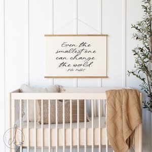 Op de afbeelding: Een witte canvasprint met zwarte tekst die luidt: "Zelfs de kleinste kan de wereld veranderen. - Peter Rabbit." De print hangt aan een witte muur boven een wit wiegje met een bruin dekentje en kussens.