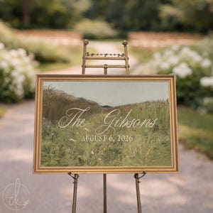 May include: A rectangular sign in a gold frame resting on a metal easel. The sign features a landscape painting of a grassy field with wildflowers and the white text "The Gibsons" in script and "AUGUST 6, 2026" in a serif font below it.