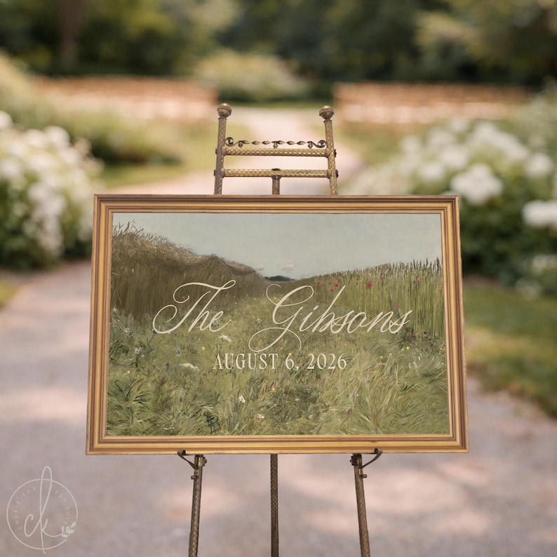 May include: A rectangular sign in a gold frame resting on a metal easel. The sign features a landscape painting of a grassy field with wildflowers and the white text "The Gibsons" in script and "AUGUST 6, 2026" in a serif font below it.