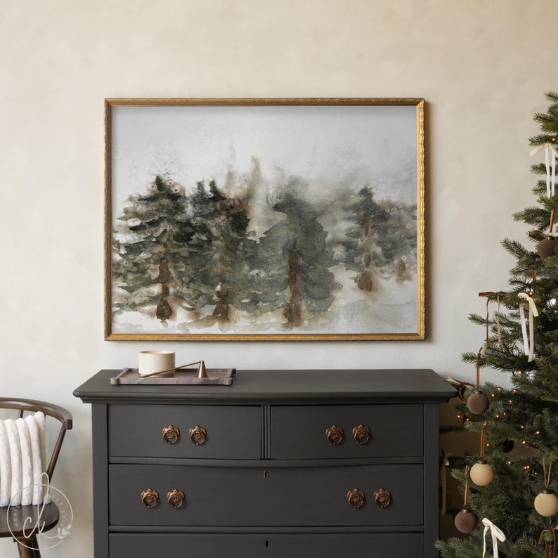 May include: A framed watercolor painting depicts a misty forest scene with tall evergreen trees in shades of green and brown. The artwork is encased in a gold-colored frame and hangs above a dark gray dresser. A Christmas tree is visible on the right.