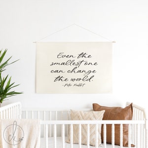 Op de afbeelding: Een witte canvas wandhanger met de quote "Zelfs de kleinste kan de wereld veranderen! - Peter Rabbit" in zwarte, cursieve letters.