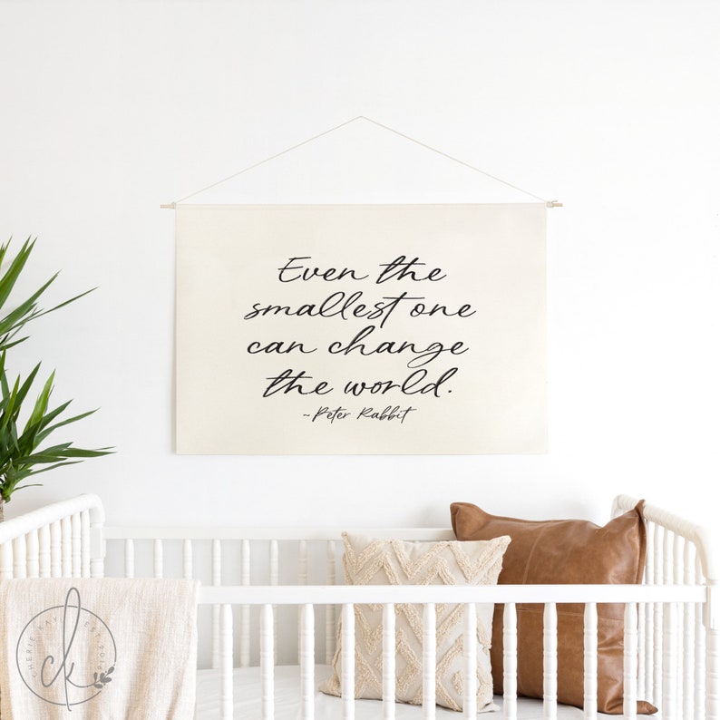 Op de afbeelding: Een witte canvas wandhanger met de quote "Zelfs de kleinste kan de wereld veranderen! - Peter Rabbit" in zwarte, cursieve letters.
