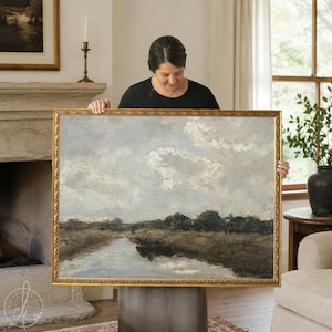 May include: A landscape oil painting in a gold ornate frame depicting a marshy river scene under a cloudy sky. The artwork features muted tones of brown, grey, and white, showcasing a calm waterway flowing through dark green vegetation.