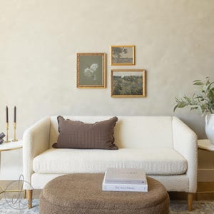May include: A cream-coloured textured sofa with a brown checkered rectangular pillow, a brown round ottoman, and two stacked books titled The Garden Journal and The Art of Home. Three gold-framed pictures hang on the wall above the sofa.