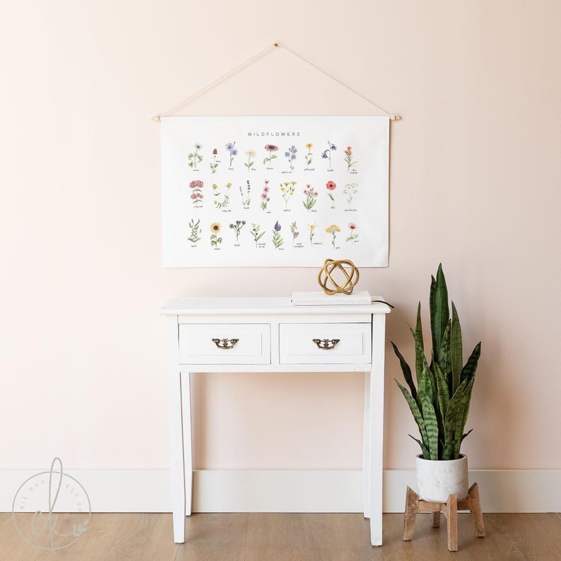 May include: A white console table with two drawers and brass hardware is positioned beneath a wall hanging that reads "Wildflowers." A gold decorative sphere and a potted snake plant on a wooden stand complete the scene.