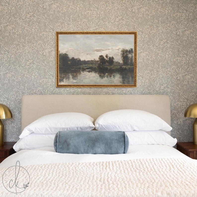 May include: A bedroom scene featuring a bed with white bedding, pillows, and a blue bolster pillow. Above the bed hangs a framed landscape painting with a gold frame. The wallpaper has a subtle floral pattern.