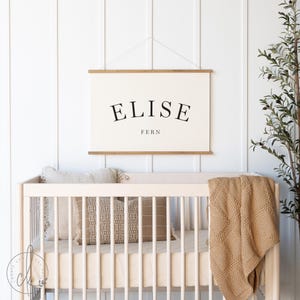 May include: A rectangular wall hanging featuring a cream-colored canvas with the name ELISE printed in a large, black, arched serif font, with the word FERN printed in a smaller, straight black font below, suspended by two wooden rods.