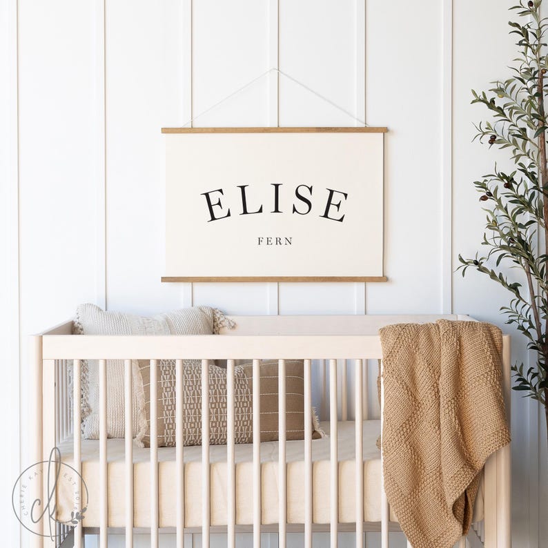 May include: A rectangular wall hanging featuring a cream-colored canvas with the name ELISE printed in a large, black, arched serif font, with the word FERN printed in a smaller, straight black font below, suspended by two wooden rods.