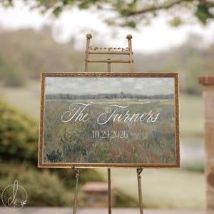 May include: A rectangular sign in a gold ornate frame featuring a landscape painting of a field with the white text The Turners and the date 10.29.2026, displayed on a gold metal easel stand. The sign has a muted green and brown color palette.