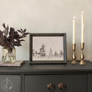 May include: A framed black and white print of a snowy forest scene sits on a dark gray dresser. Two lit, twisted ivory candles stand in gold candle holders. A glass vase with dark purple leaves is on the left.