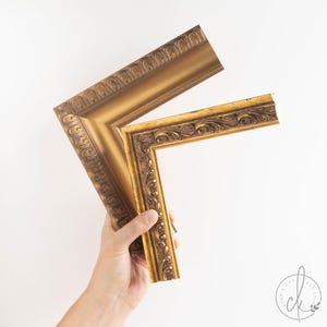 May include: Two sections of a gold picture frame with detailed, ornate carvings. The frame has a classic, vintage style and a warm, metallic finish. The frame is held against a white background, showcasing its decorative elements.