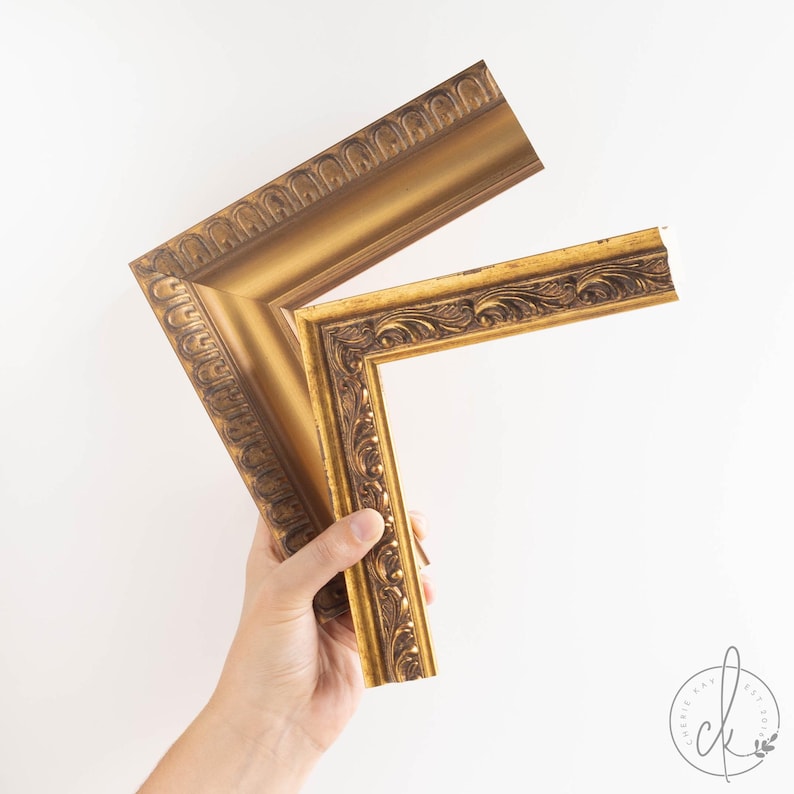 May include: Two sections of a gold picture frame with detailed, ornate carvings. The frame has a classic, vintage style and a warm, metallic finish. The frame is held against a white background, showcasing its decorative elements.