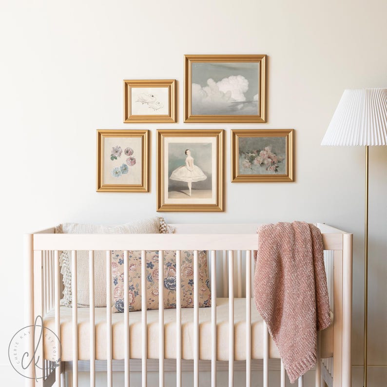 A nursery with a light wooden crib, soft bedding, and a pink blanket. Above the crib, a gallery wall features five gold-framed art prints, including florals, a ballet dancer, and clouds. A white floor lamp stands nearby.