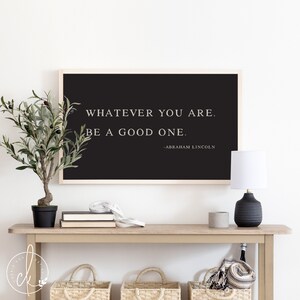 May include: A framed black print with the quote "WHATEVER YOU ARE. BE A GOOD ONE." by Abraham Lincoln. The print is displayed on a light wood console table with a small potted plant, books, and a lamp.