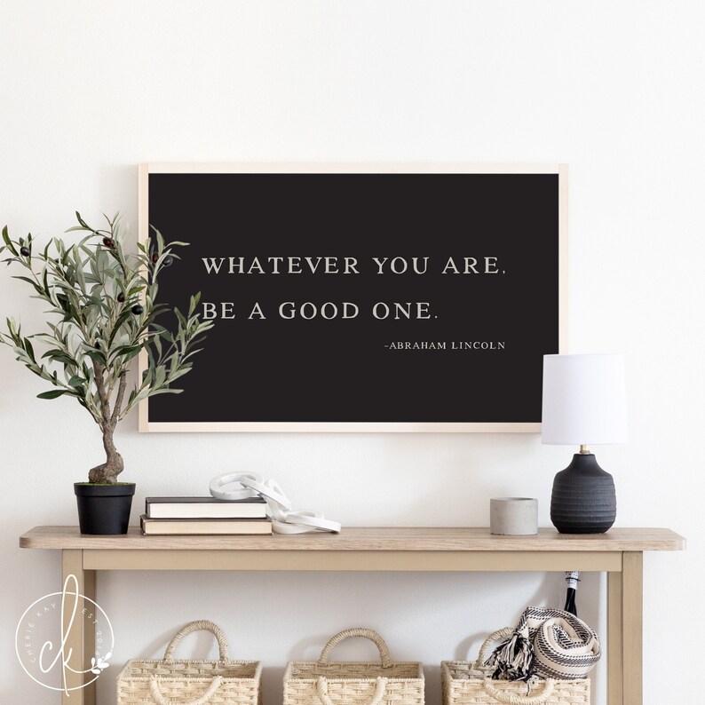 May include: A framed black print with the quote "WHATEVER YOU ARE. BE A GOOD ONE." by Abraham Lincoln. The print is displayed on a light wood console table with a small potted plant, books, and a lamp.