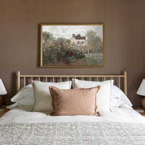 May include: A bedroom with a landscape painting above a bed. The painting shows a house and garden. The bed has white pillows, a peach-coloured cushion, and a patterned bedspread. The headboard is made of light-coloured wood.