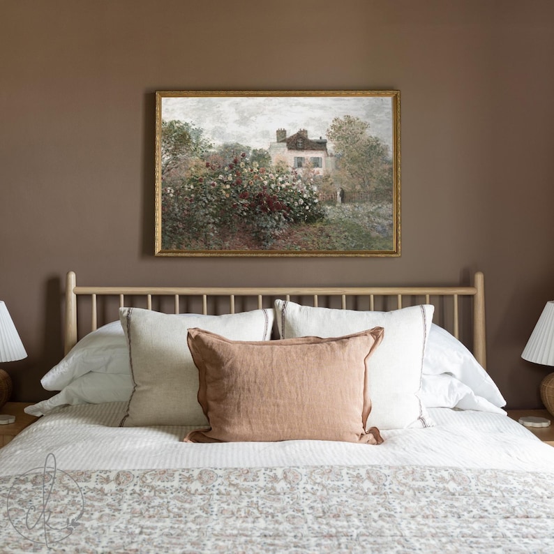 May include: A bedroom with a landscape painting above a bed. The painting shows a house and garden. The bed has white pillows, a peach-coloured cushion, and a patterned bedspread. The headboard is made of light-coloured wood.