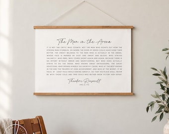May include: A white canvas wall hanging featuring the text of The Man in the Arena by Theodore Roosevelt, dated April 23, 1910. The canvas is secured between two horizontal wooden slats and suspended by a thin white string for wall display.