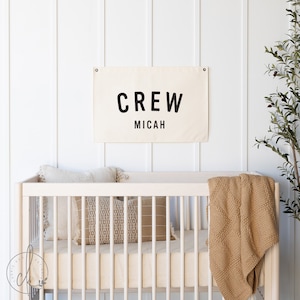 May include: A white canvas banner with black text that reads "CREW Micah" hanging on a white wall above a white crib with a tan blanket and pillows.