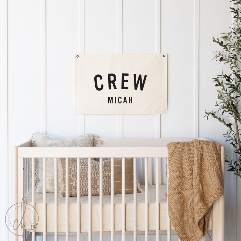 May include: A white canvas banner with black text that reads "CREW Micah" hanging on a white wall above a white crib with a tan blanket and pillows.