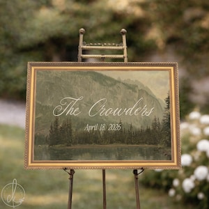 May include: A rectangular sign in a gold frame featuring a landscape painting of a mountain range, a forest, and a lake. The text "The Crowders" is printed in white script above the date "April 18, 2026" in a simple white font on the image.