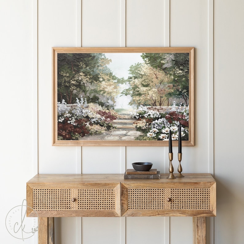 Puede incluir: Una pintura enmarcada de un camino de jard&iacute;n que conduce a trav&eacute;s de un exuberante bosque verde con flores blancas a cada lado. La pintura est&aacute; colgada en una pared blanca sobre una mesa consola de madera con dos cajones.