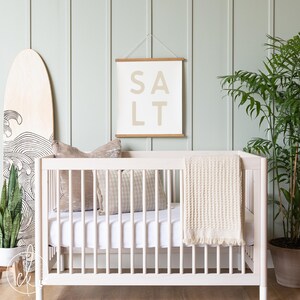 May include: A light-colored wooden crib with a white mattress, a beige waffle-knit blanket with fringe, and two decorative pillows. Above the crib hangs a white wall hanging with wooden rails and the word SALT printed in large beige letters.