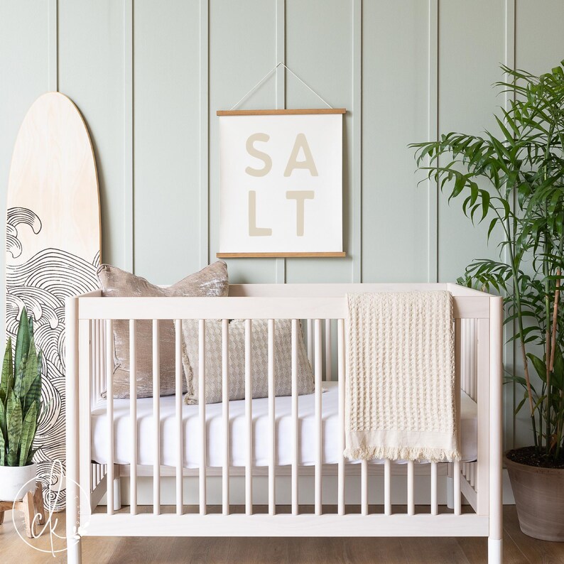 May include: A light-colored wooden crib with a white mattress, a beige waffle-knit blanket with fringe, and two decorative pillows. Above the crib hangs a white wall hanging with wooden rails and the word SALT printed in large beige letters.