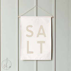 May include: A rectangular off-white fabric wall hanging featuring the word SALT printed in large, bold, beige sans-serif capital letters. The banner is suspended from a thin wooden dowel with a white string attached to both ends for hanging.