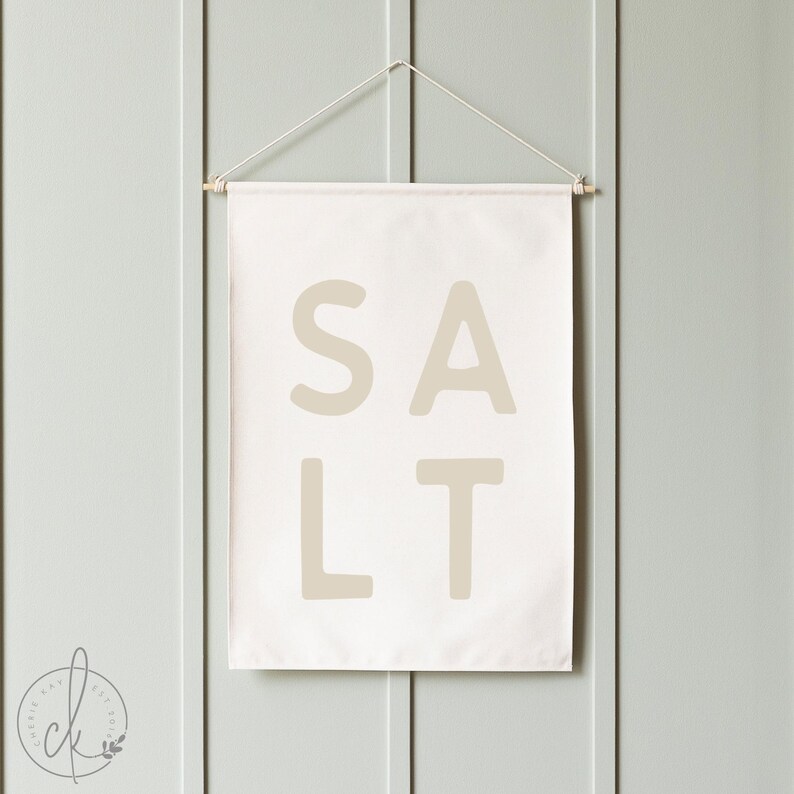 May include: A rectangular off-white fabric wall hanging featuring the word SALT printed in large, bold, beige sans-serif capital letters. The banner is suspended from a thin wooden dowel with a white string attached to both ends for hanging.
