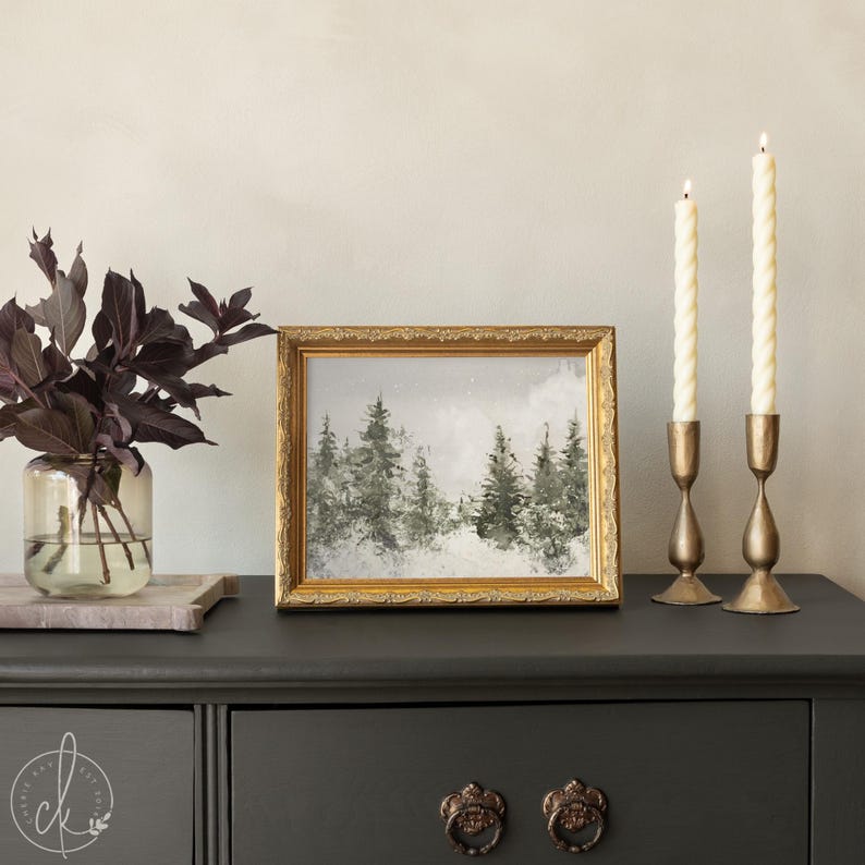 May include: A framed landscape painting featuring a forest of evergreen trees in shades of gray and white, encased in an ornate gold frame. Beside it are two cream-colored twisted taper candles in brass holders and a glass vase with dark foliage.